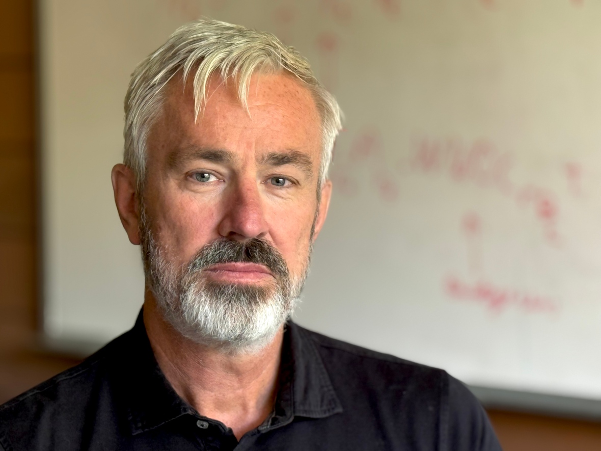 A man wearing a black polo shirt poses for a photograph inside a university classroom.